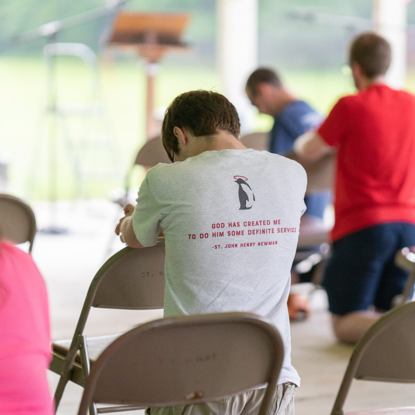 Young adult bowed in prayer with t-shirt reading "God has created me to do him some definite service." -St. John Henry Newman