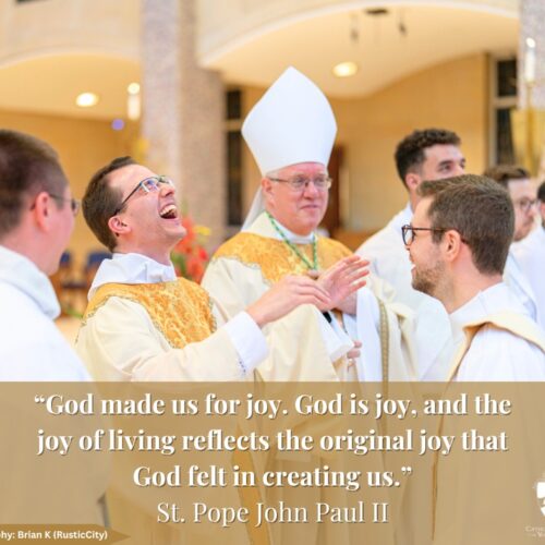 Travis Seagraves laughing with vestments on, surrounded by servers, deacons and bishop in vestments. Quote: "God made us for joy. God is joy, and the joy of living reflects the original joy that God felt in creating us." Saint Pope Johne Paul II