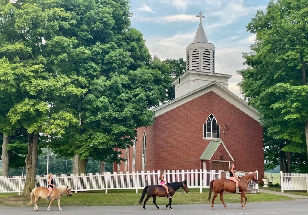 Three rides on horses in front of white picket fence and red brick church with white wooden steeple and cross