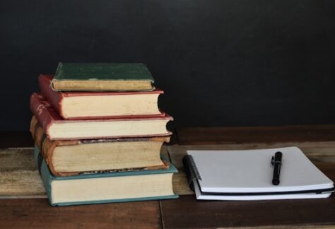 Piles of hard bound books beside an open notebook and pen