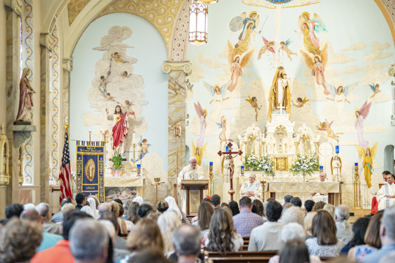 Ornate church with murals, large statues of Mary and Jesus, white marble altar, golden candlesticks, flower arrangements, and banner reading "Basilica of Our Lady of Mount Carmel, 2014"