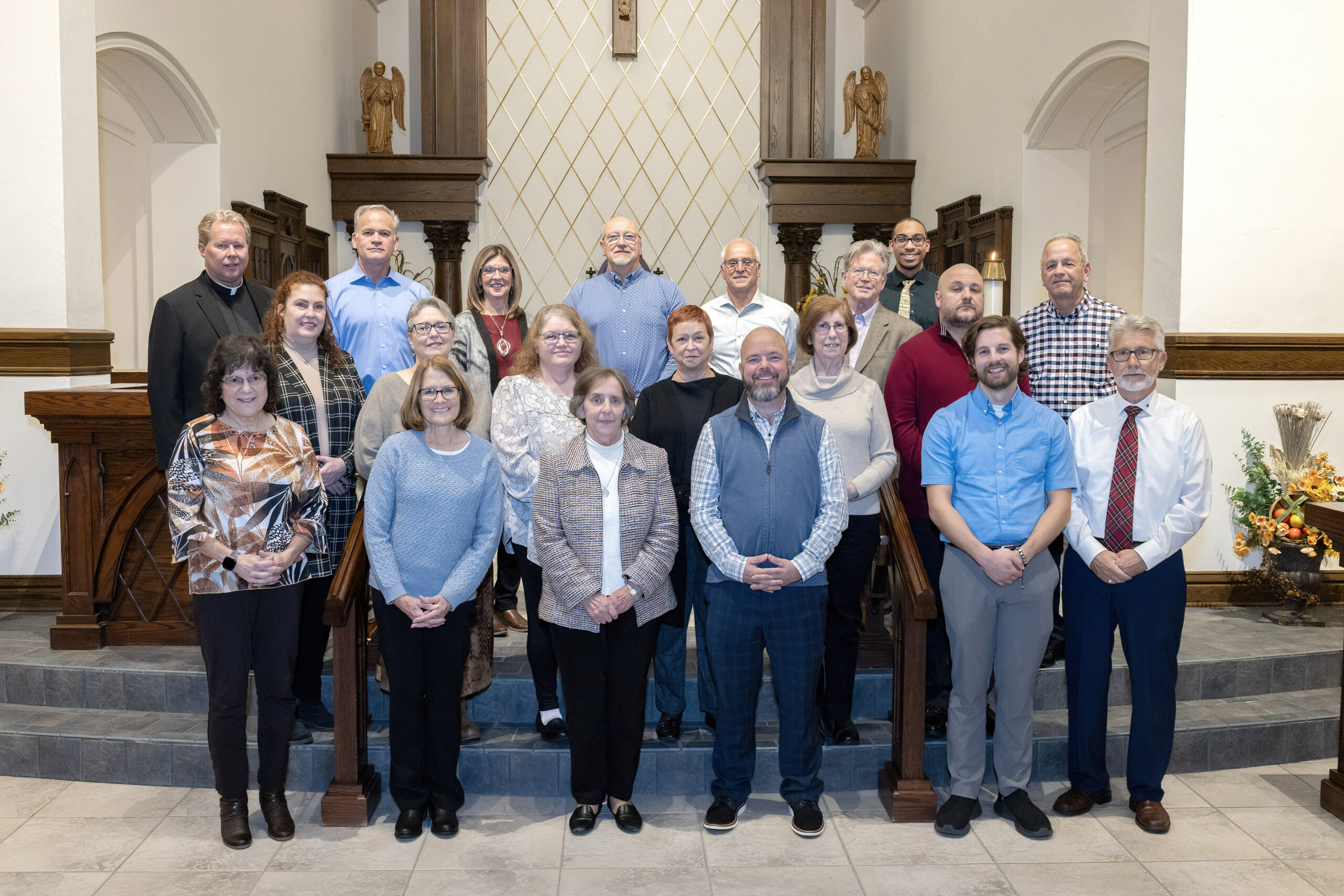 20 people lined up in three rows on steps in the sanctuary of a church, smiling