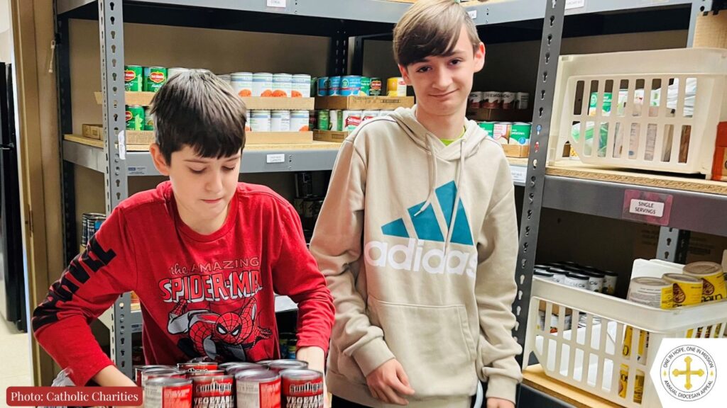 Two teens carry donations of cans in a Catholic Charities food pantry with stocked shelves