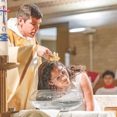 Father Frazzati in gold vestments pours water from a seashell over the head of a young woman wearing white to baptize her during Easter Vigil