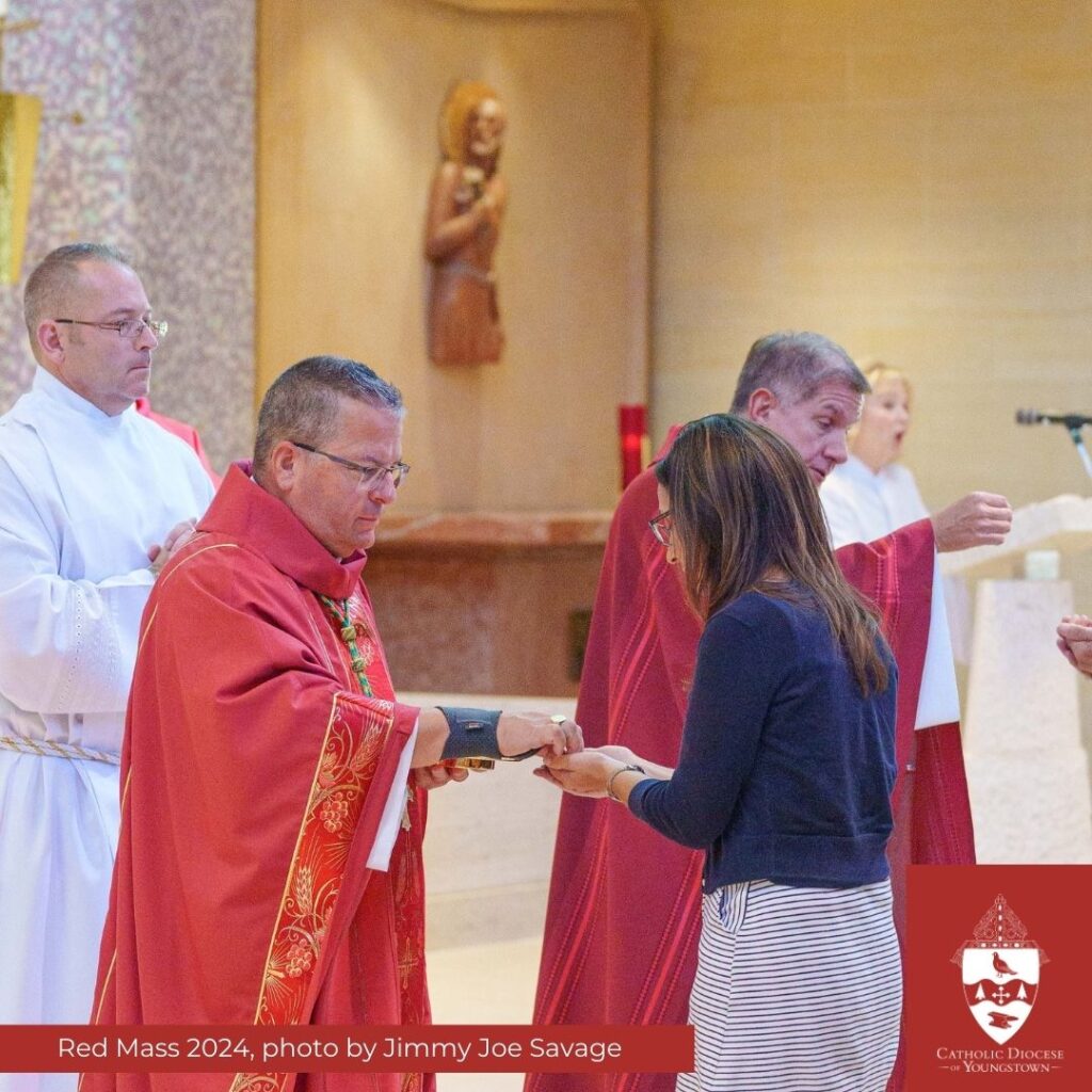 Two people in red vestments distribute Communion with a server and cantor in white robes in the background and a statue of St. Joseph on the wall