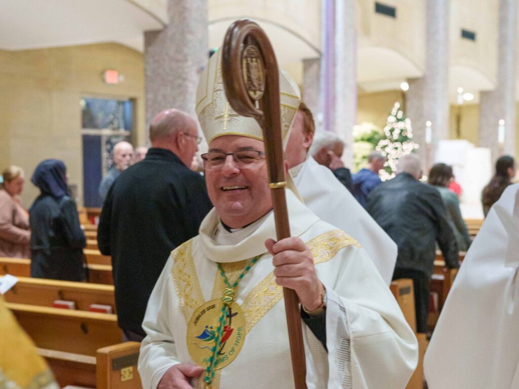 Bishop Bonnar holds his staff and wears white vestments with gold trim and the Jubilee 2025 logo that reads "Pilgrims of Hope."