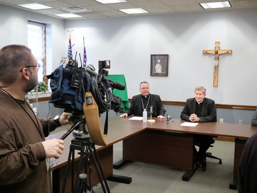 TV new camera films smiling Bishop and Vicar General with crucifix in the background