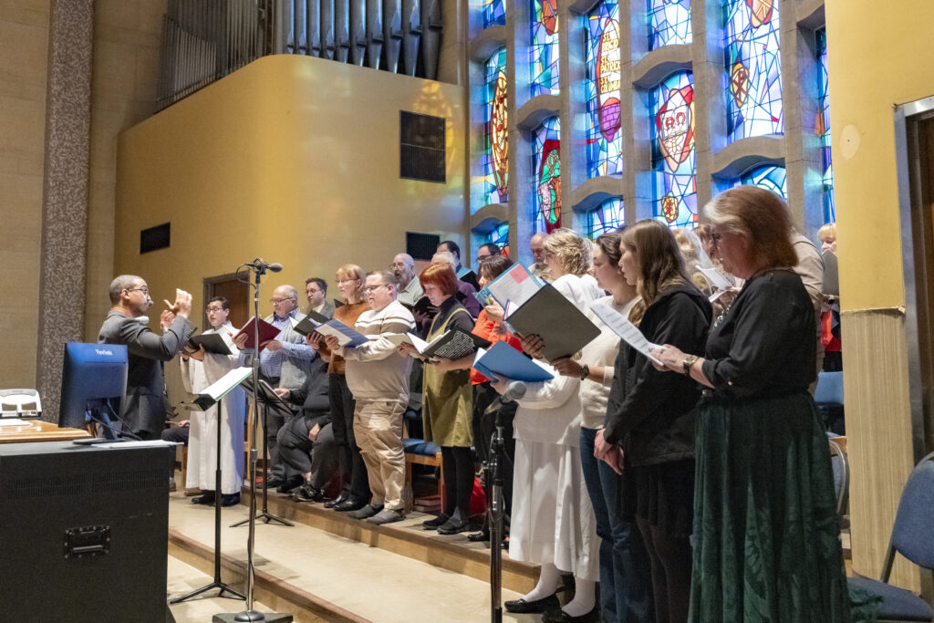 Man in suit conducts before risers full of singing choristers in front of stained glass window