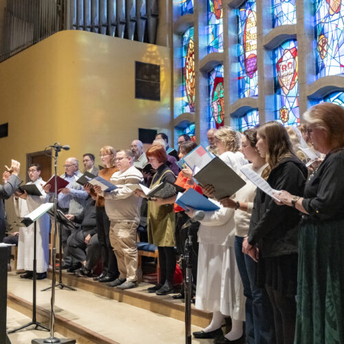 Man in suit conducts before risers full of singing choristers in front of stained glass window