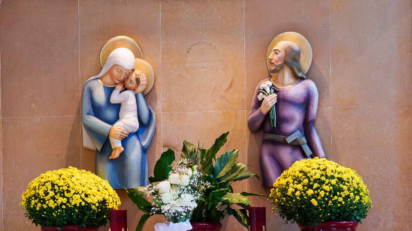 Candles lit in front of statues of the Holy Family: Jesus in Mary's arms, and Joseph gazing at them while holding woodworking tools