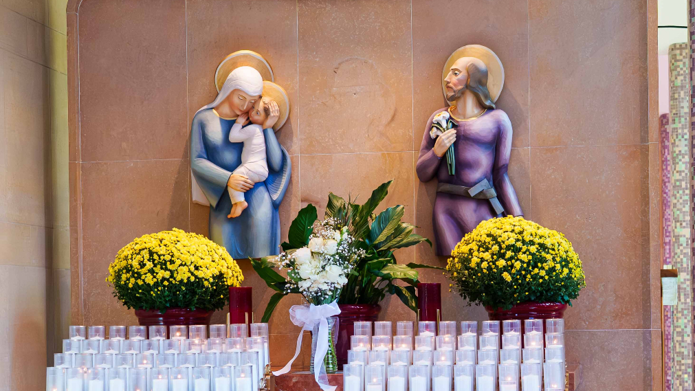 Candles lit in front of statues of the Holy Family: Jesus in Mary's arms, and Joseph gazing at them while holding woodworking tools