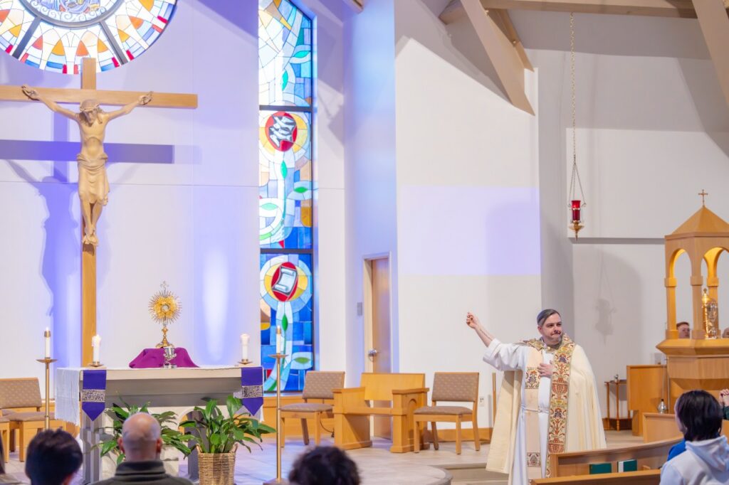 Fr. Chad Johnson in vestments stands before an altar covered in purple for Lent pointing at the large crucifix behind him.
